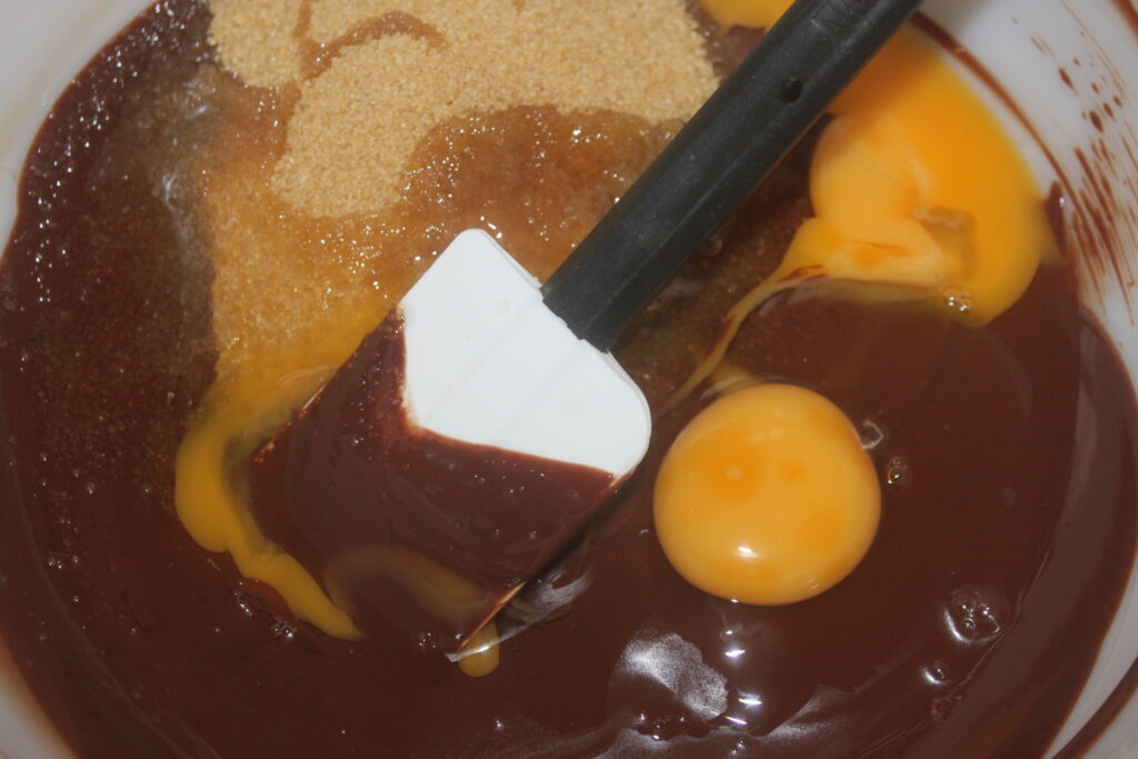 Un bol à mélanger contenant une spatule, du chocolat fondu, de la cassonade, des œufs cassés et un soupçon d'amande évoque le début d'un délicieux gâteau au chocolat avec de riches pépites de chocolat.