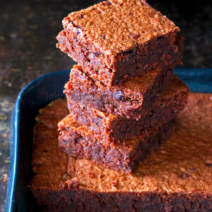 Une pile de quatre brownies au chocolat carrés repose sur une plaque de cuisson sombre, rappelant un gâteau décadent, avec des friandises plus délicieuses en arrière plan.