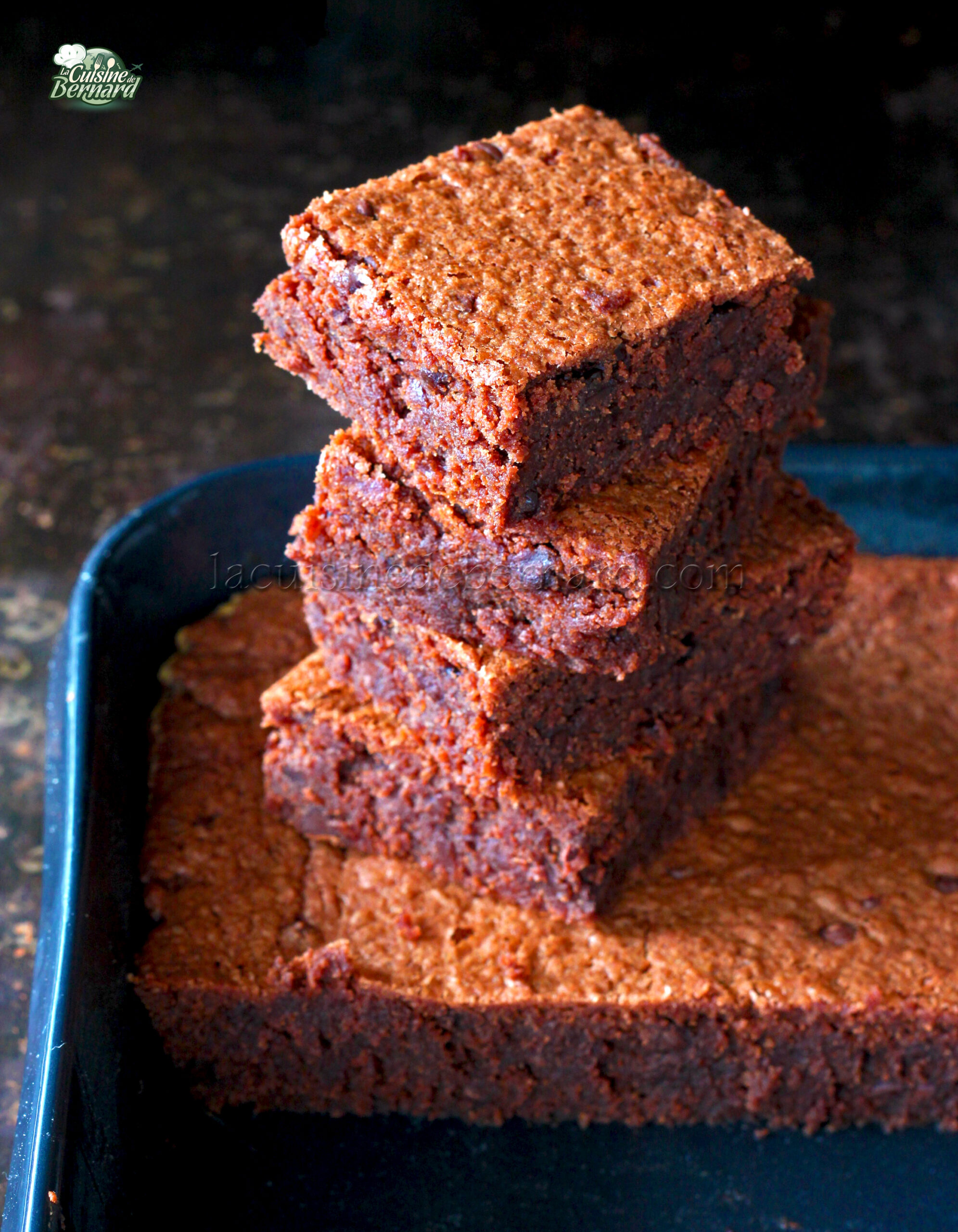 Une pile de quatre brownies au chocolat carrés repose sur une plaque de cuisson sombre, rappelant un gâteau décadent, avec des friandises plus délicieuses en arrière plan.