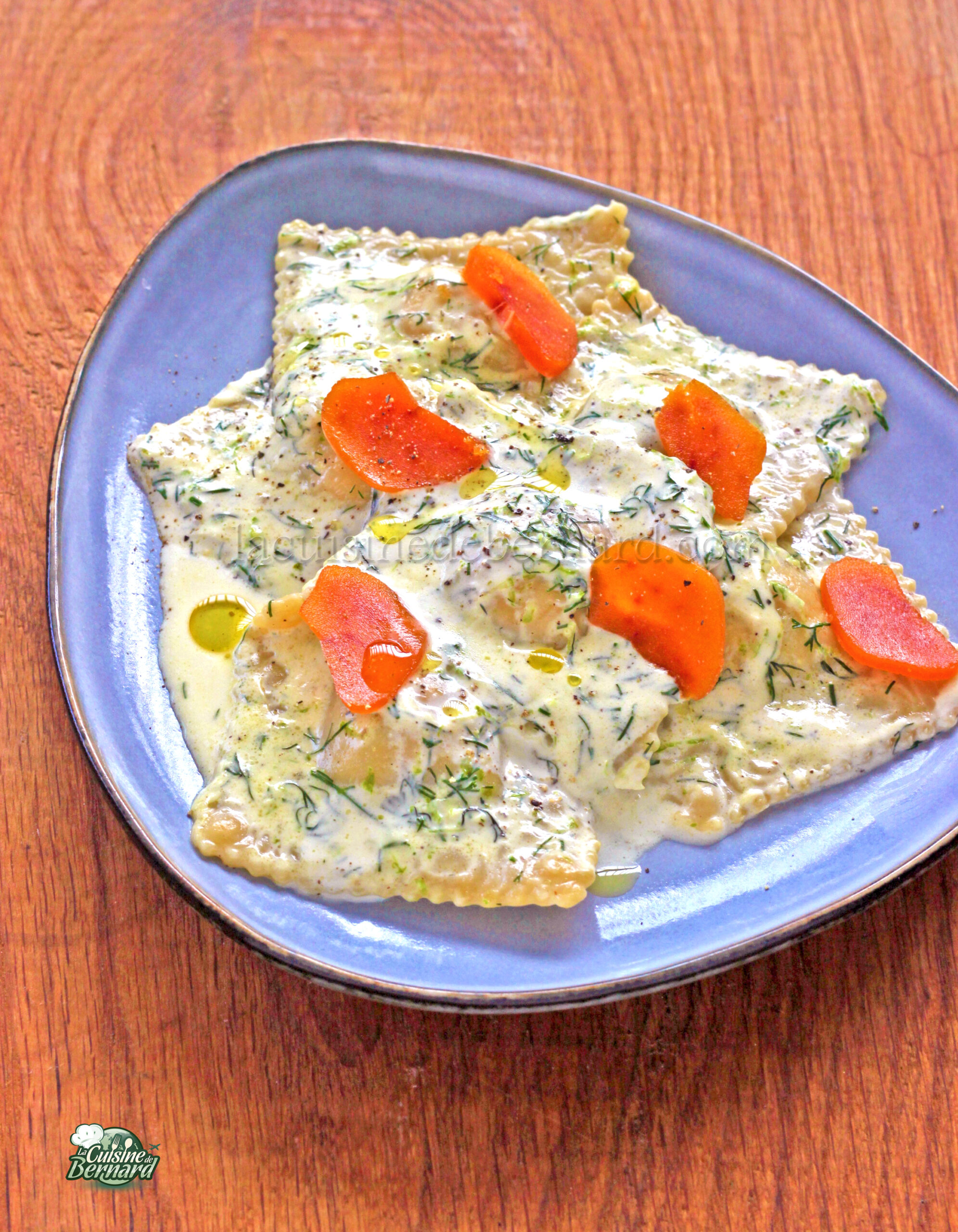Une assiette bleue contient des raviolis recouverts d'une sauce crémeuse à l'aneth, garnis de tranches de carottes orange et d'herbes, agrémentées d'une pointe de citron vert, sur une table en bois.
