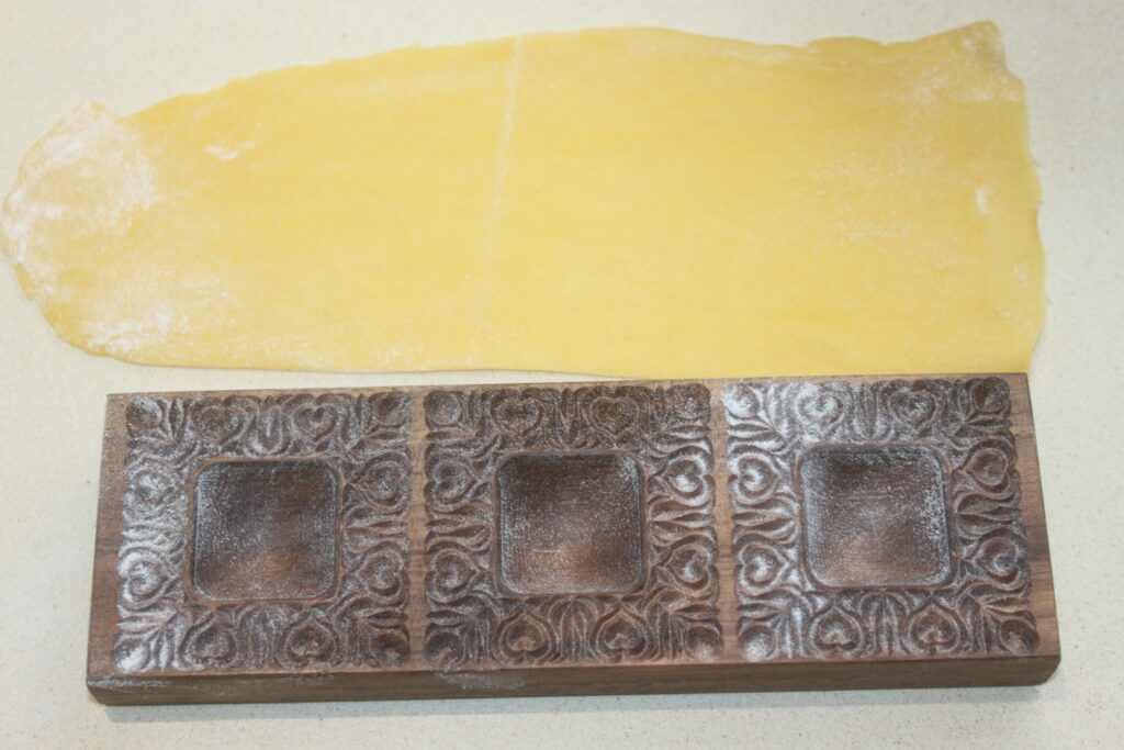 Un moule à raviolis en bois aux motifs décoratifs repose à côté d'une feuille de pâte à pâtes roulée, prêt à confectionner de délicieux raviolis à la ricotta sur une surface claire.
