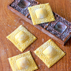 Quatre morceaux de raviolis carrés aux motifs décoratifs sur une surface en bois, à côté d'un moule en bois sculpté, sont soigneusement remplis de ricotta, ajoutant une texture crémeuse qui complète leur design délicat.