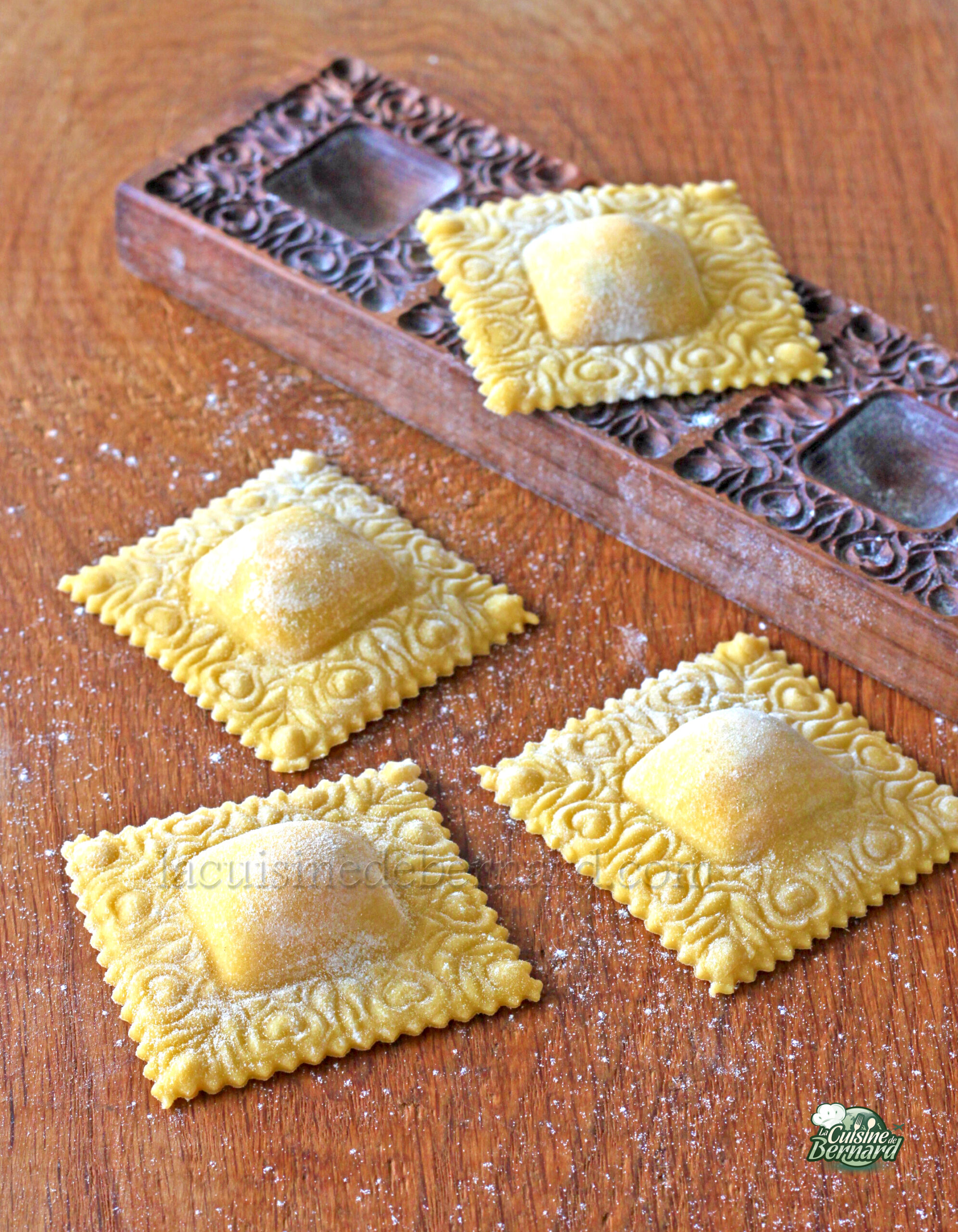 Quatre morceaux carrés de pâte à raviolis décorative sont placés près d'un moule à pâtes en bois sur une surface farinée, prêts à être transformés en exquis raviolis saumon.