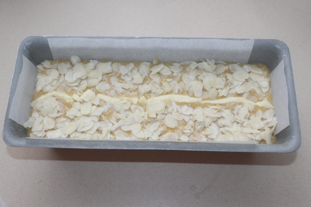 Un moule à pain rectangulaire recouvert de papier sulfurisé rempli de pâte à gâteau, garni de tranches d'amandes, prêt à être cuit.