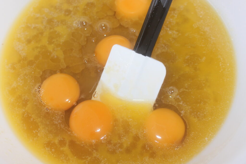 Un bol à mélanger contenant des œufs et du beurre fondu est prêt pour votre prochaine aventure culinaire, avec une spatule partiellement immergée dans le mélange. Parfaite pour préparer un délicieux gâteau, cette configuration garantit que chaque étape de votre mégamande est fluide et agréable.