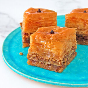Gros plan de trois morceaux de baklava en couches avec noix sur une assiette turquoise. Chaque pièce est surmontée d'un clou de girofle, mettant en valeur les riches traditions des spécialités grecques.