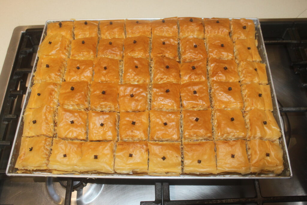 Un plateau de baklava fraîchement sorti du four avec des couches de pâte feuilletée dorées, chaque morceau surmonté d'un clou de girofle et parsemé de noix finement hachées, placé sur une cuisinière.