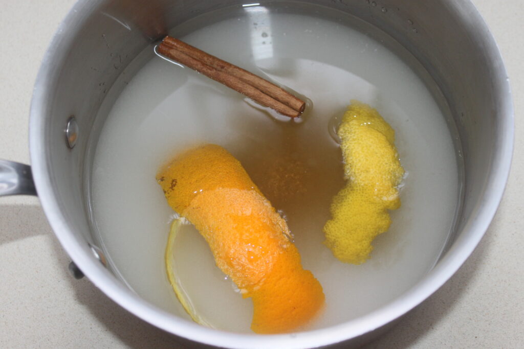Une casserole avec de l'eau, des zestes d'orange et de citron, un bâton de cannelle et un soupçon de noix.