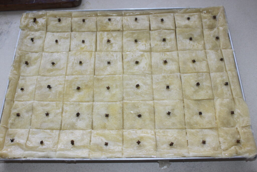 Un plateau de baklava non cuit, chaque morceau carré surmonté d'un clou de girofle et prêt à être cuit, met en valeur les riches saveurs de noix de ce dessert grec classique.