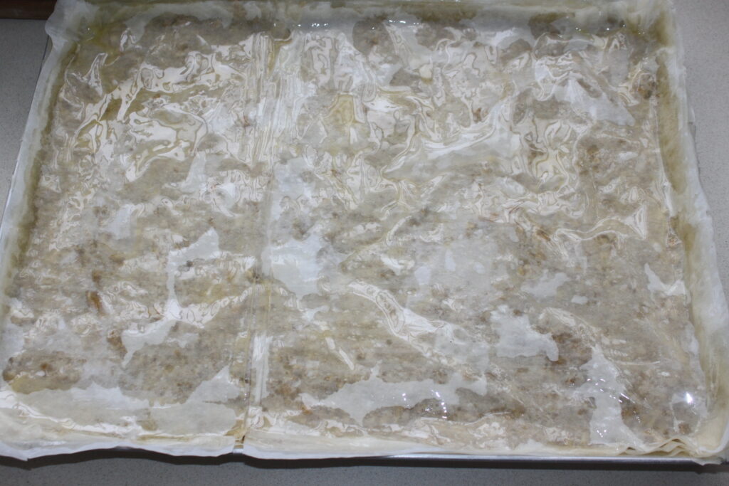 Un plateau rectangulaire enveloppé de plastique rempli d'un mélange qui semble aqueux et grumeleux, reposant sur une surface de couleur claire, rappelant la texture du Baklava traditionnel.