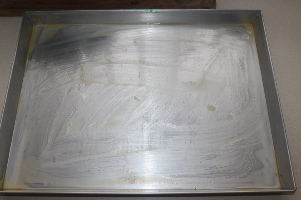 Une plaque à pâtisserie en métal est présentée à moitié recouverte d'une fine couche de beurre, prête à créer de délicieux baklava.