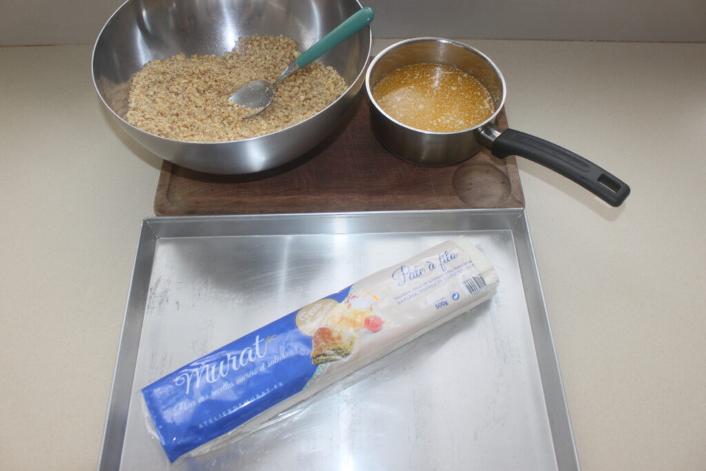 Un bol d'ingrédients secs mélangés avec une spatule, une casserole de beurre fondu et un paquet de pâte à pâte à filo sur une planche à découper sur une plaque à pâtisserie : des essentiels parfaits pour préparer de délicieux Baklava riches en noix comme le voulaient les Grecs.