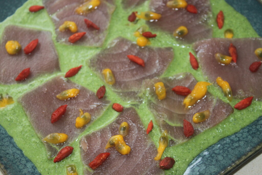 Une assiette de thon cru tranché finement sur une sauce verte, garnie de baies de goji et de vinaigrette jaune.