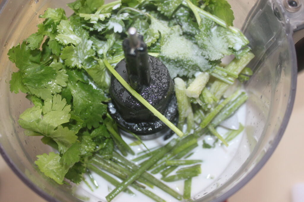 Gros plan de divers légumes verts et herbes feuillues dans un robot culinaire, ressemblant au mélange vibrant utilisé dans un ceviche rafraîchissant, avec une petite quantité de liquide au fond.