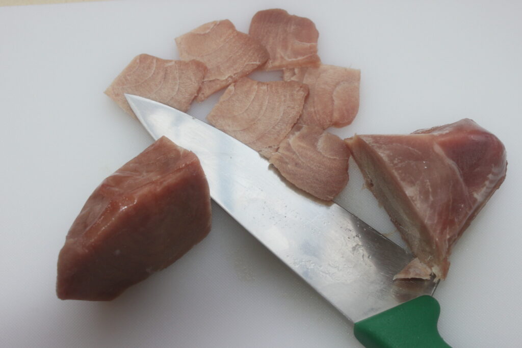 Des tranches et des morceaux de viande crue reposent sur une planche à découper blanche, avec un couteau à manche vert partiellement traversant un seul morceau, parfait pour préparer un ceviche frais.