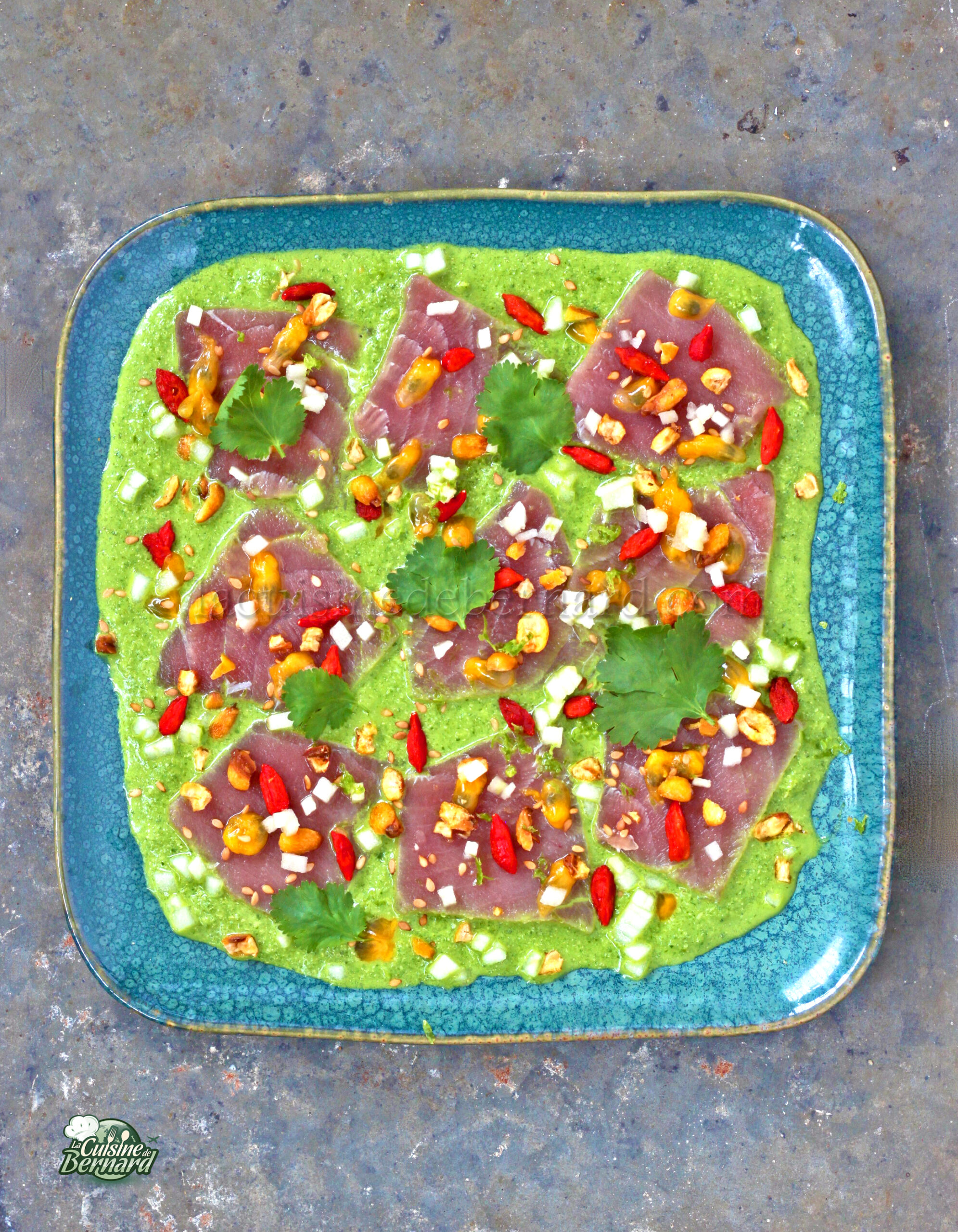 Une assiette carrée bleue présente des tranches de thon cru sur une sauce en purée verte, garnie de piments rouges, de noix et de feuilles de coriandre, évoquant une touche fusion au ceviche traditionnel.