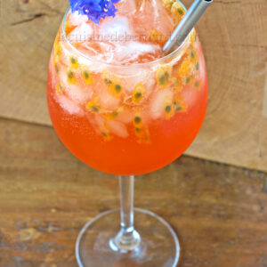 Un spritz rouge orange vibrant avec de la glace, des graines de fruits de la passion, une paille métallique et une garniture de fleurs bleues dans un grand verre à vin sur une surface en bois, évoquant l'ambiance de la plage.
