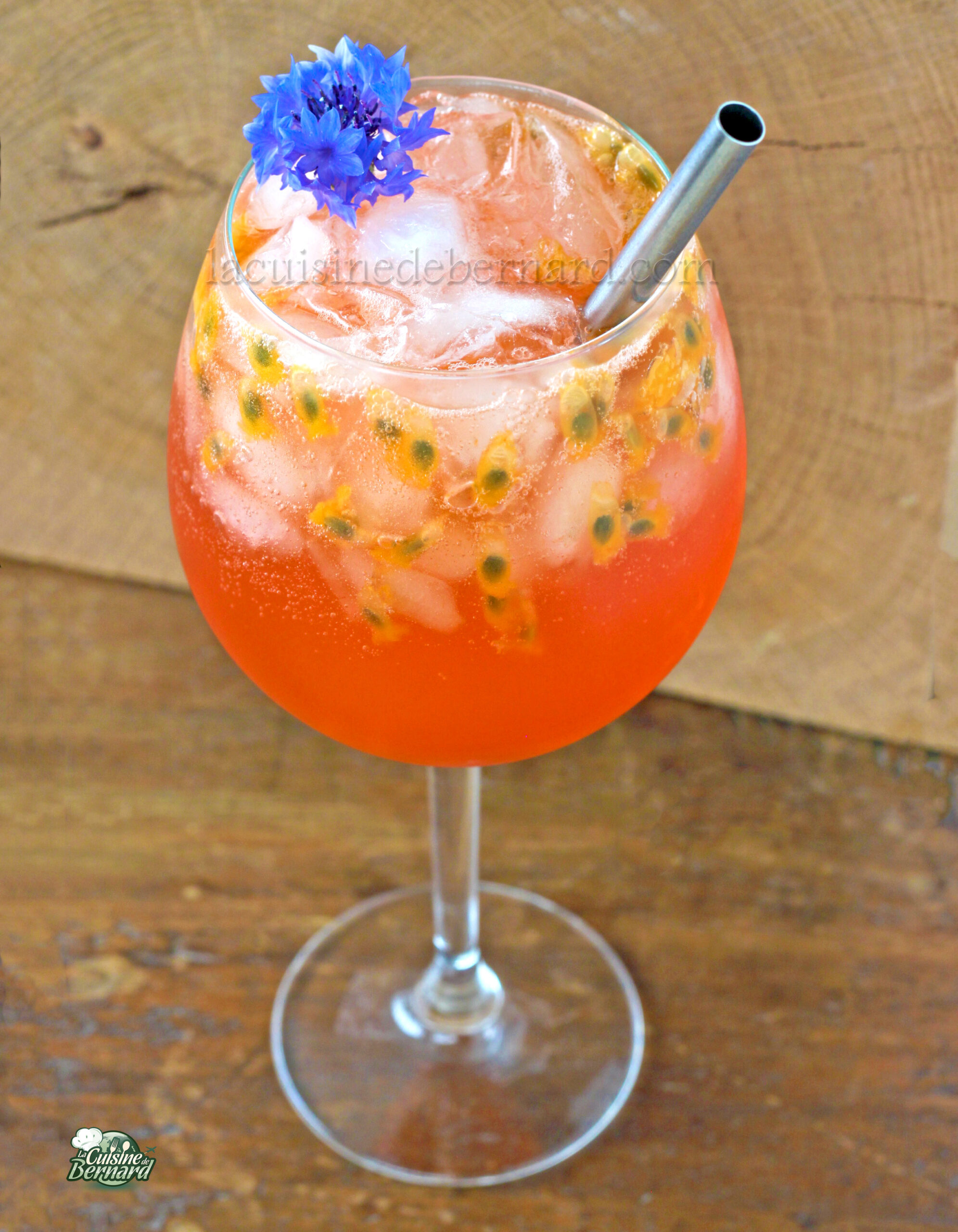 Un spritz rouge orange vibrant avec de la glace, des graines de fruits de la passion, une paille métallique et une garniture de fleurs bleues dans un grand verre à vin sur une surface en bois, évoquant l'ambiance de la plage.