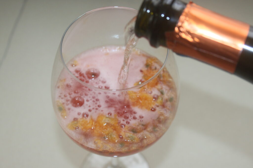 Un verre de vin rempli d'un spritz pétillant provenant d'une bouteille. La boisson a un dessus mousseux avec quelques morceaux de fruits visibles, parfait pour déguster à la plage.