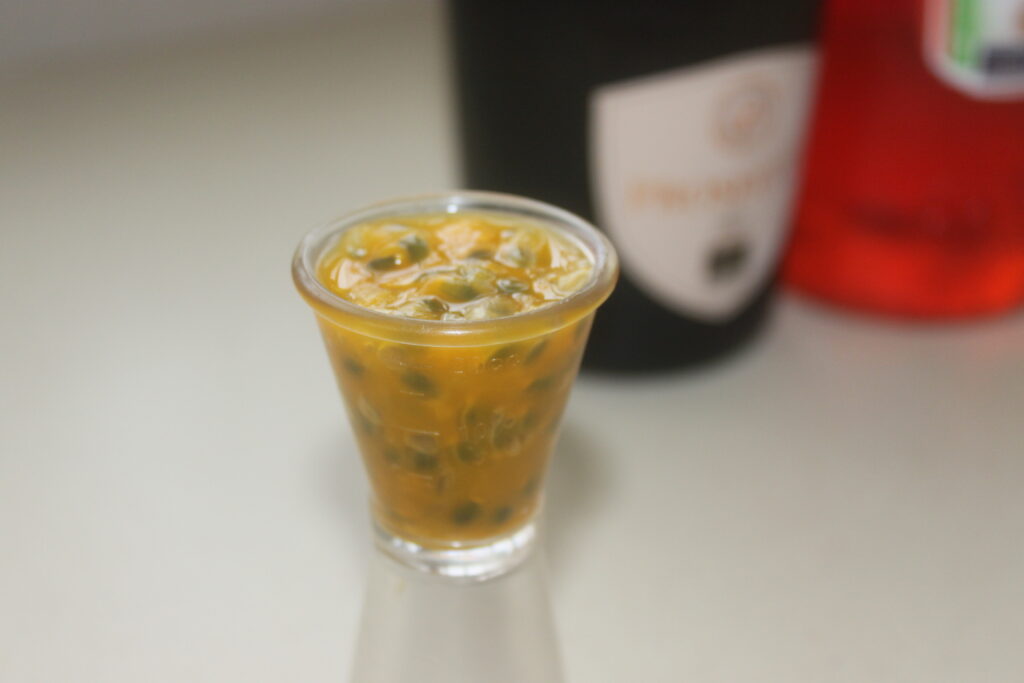 Un petit verre rempli de pulpe de fruit de la passion, évoquant un spritz de délice tropical, posé sur un comptoir avec des bouteilles floues en arrière plan, rappelant un bar de plage venteux.