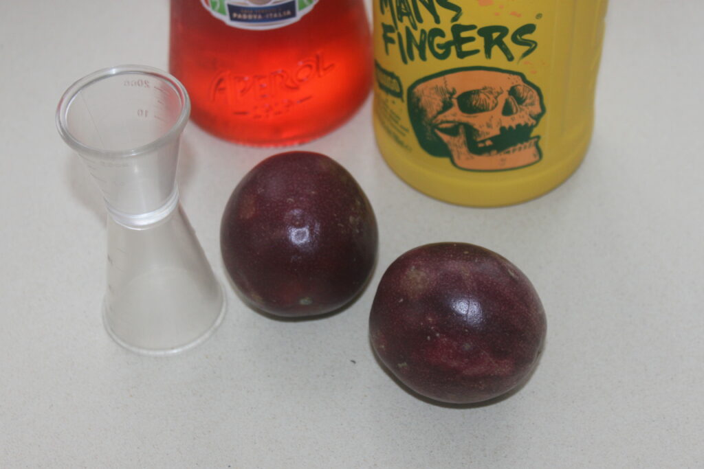 Deux fruits de la passion violet foncé sont placés sur une surface blanche à côté d'un petit récipient doseur en plastique transparent, d'une bouteille d'« Aperol » et d'une bouteille étiquetée « Man's Fingers », prêts à préparer un spritz rafraîchissant en bord de plage.