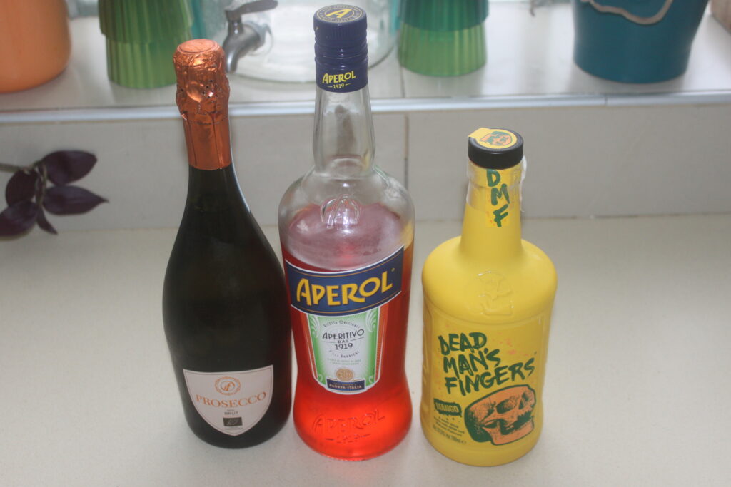 Trois bouteilles sont exposées sur un comptoir : une bouteille de prosecco avec du papier d'aluminium marron, une bouteille d'Aperol remplie de liquide rouge et une bouteille Dead Man's Fingers arborant un motif de crâne jaune vif. Parfait pour préparer le spritz de plage idéal.
