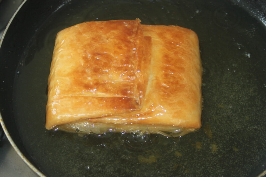 Une pâtisserie carrée à la surface dorée, fourrée de Feta croustillante, est frite dans une poêle avec de l'huile.