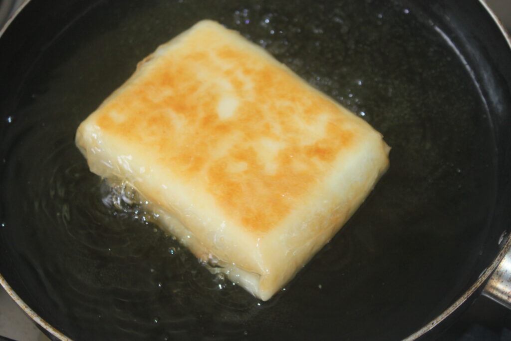 Un morceau rectangulaire de tofu est frit dans une poêle avec de l'huile, présentant une surface brun doré, et est garni de feta émiettée.