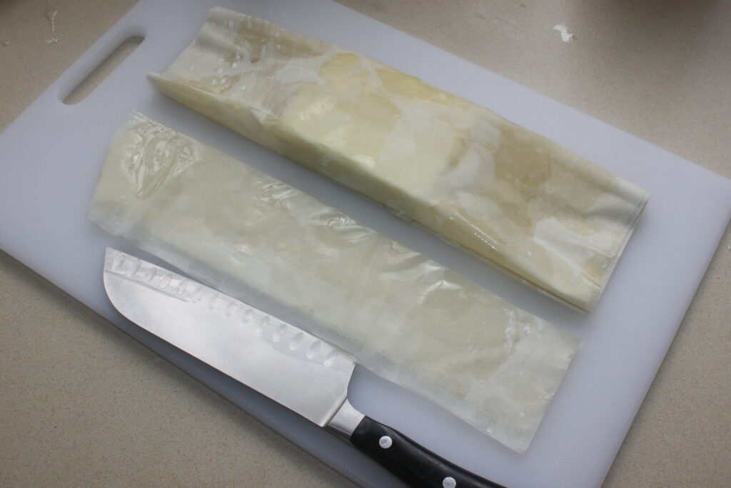 Deux enveloppes de cire pliées avec un couteau de chef sur une planche à découper blanche, faisant allusion à un prochain plat composé de Feta croustillante et de pistache.