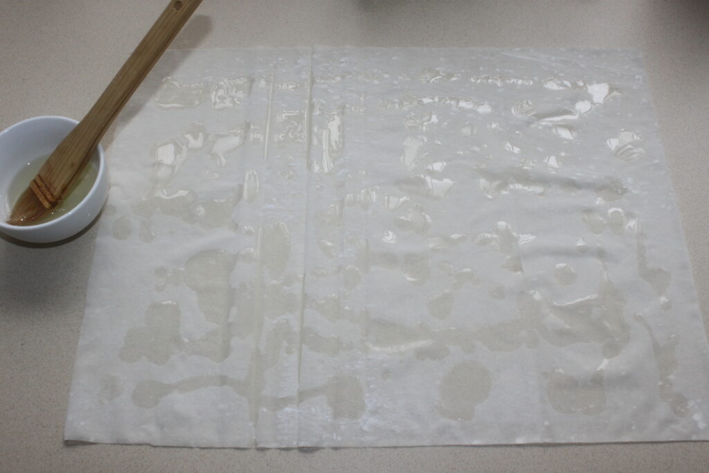 Une feuille de papier de riz humide et translucide repose à plat sur un comptoir à côté d'un bol, un pinceau probablement utilisé pour appliquer de l'eau et un plat de feta croustillante avec des notes de miel et de pistache.