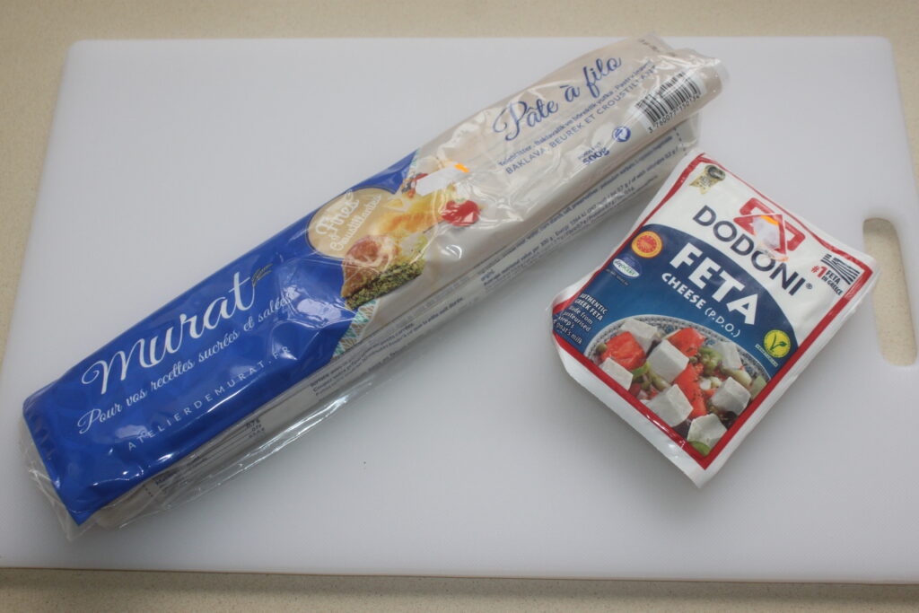 Un paquet de pâte filo de marque Murat côtoie un paquet de feta Dodoni, tous deux posés sur une planche à découper blanche, prêts à être transformés en une délicieuse Feta croustillante avec un filet de miel.