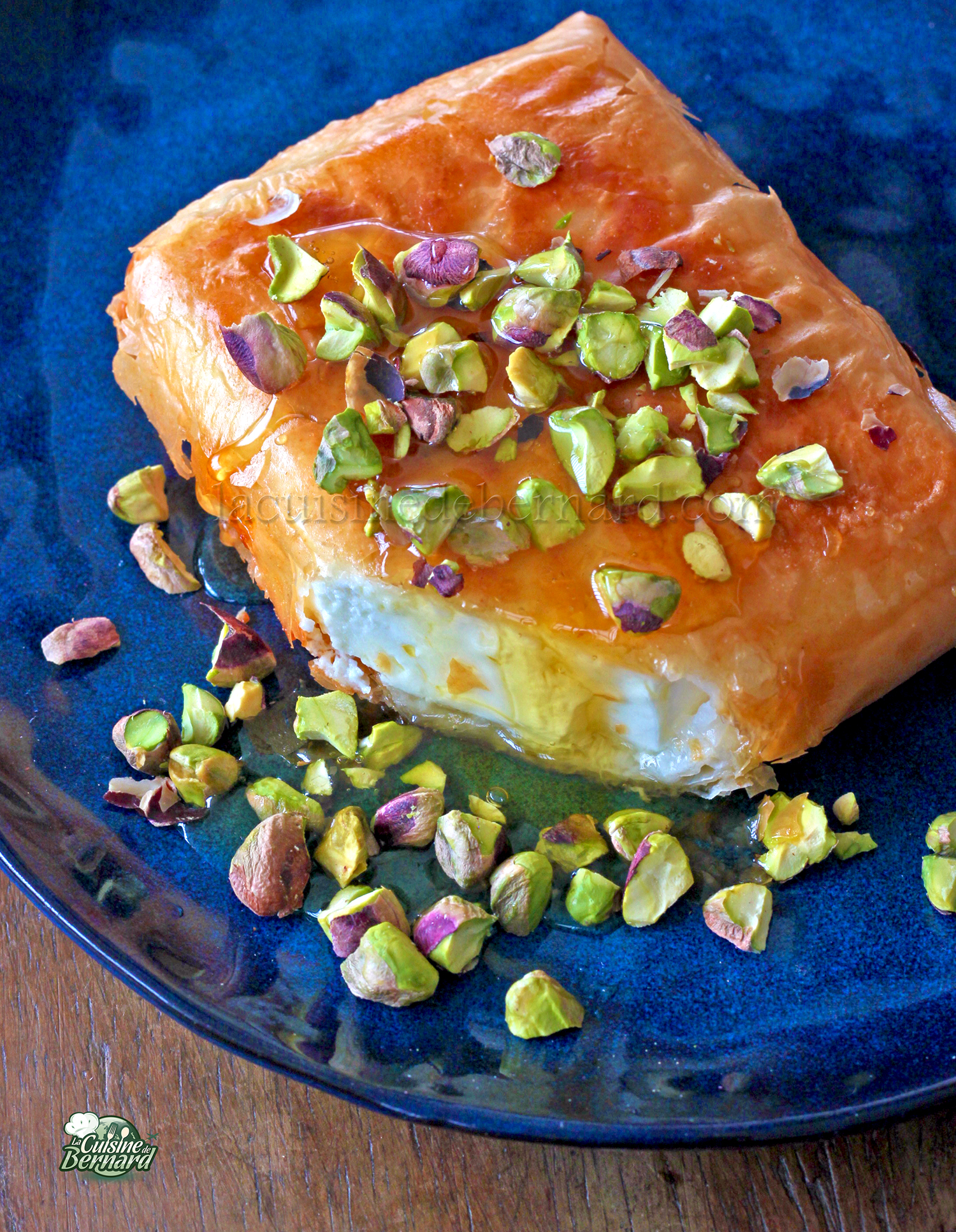 Une tranche de feta croustillante enveloppée dans de la pâte phyllo, garnie de miel et de pistache hachée, servie sur une assiette bleue.