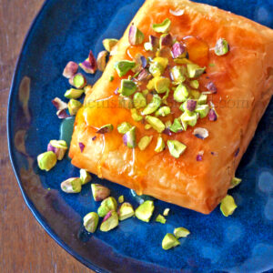 Un dessert composé d'une pâtisserie bien dorée, arrosée de miel et garnie de pistache hachée, est servi sur une assiette bleue.