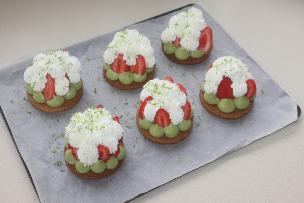 Six Tartelettes fraîches à base de crème verte, fraises fraîches et chantilly, garnies de zestes de citron vert, disposées sur une plaque à pâtisserie tapissée de papier sulfurisé.