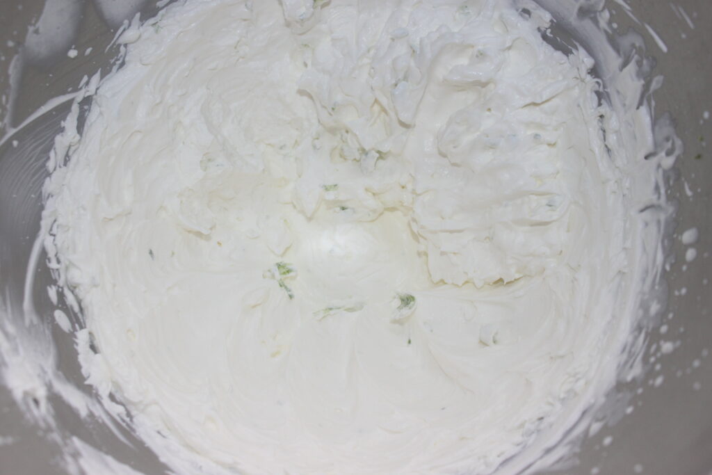 Un bol contient de la crème blanche fouettée épaisse avec quelques taches vertes, probablement un mélange de chantilly mascarpone ou un produit similaire.