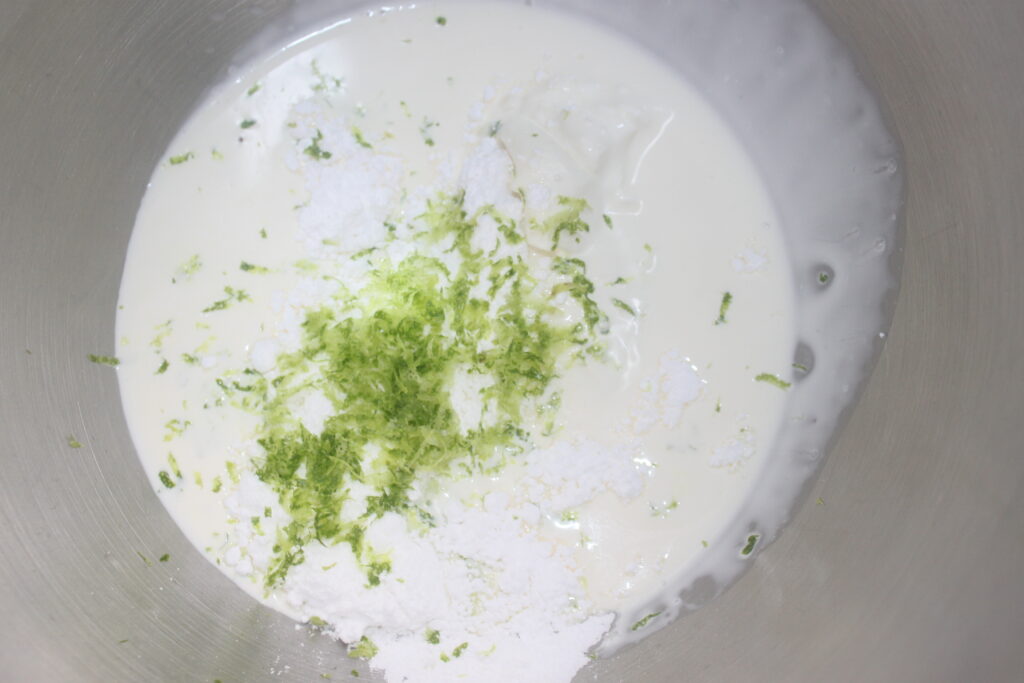 Un bol à mélanger contenant de la crème, du sucre en poudre, du zeste vert (probablement du citron vert) ainsi qu'un soupçon de chocolat blanc, prêts à être mélangés dans une délicieuse chantilly mascarpone pour tartelettes fraîches.