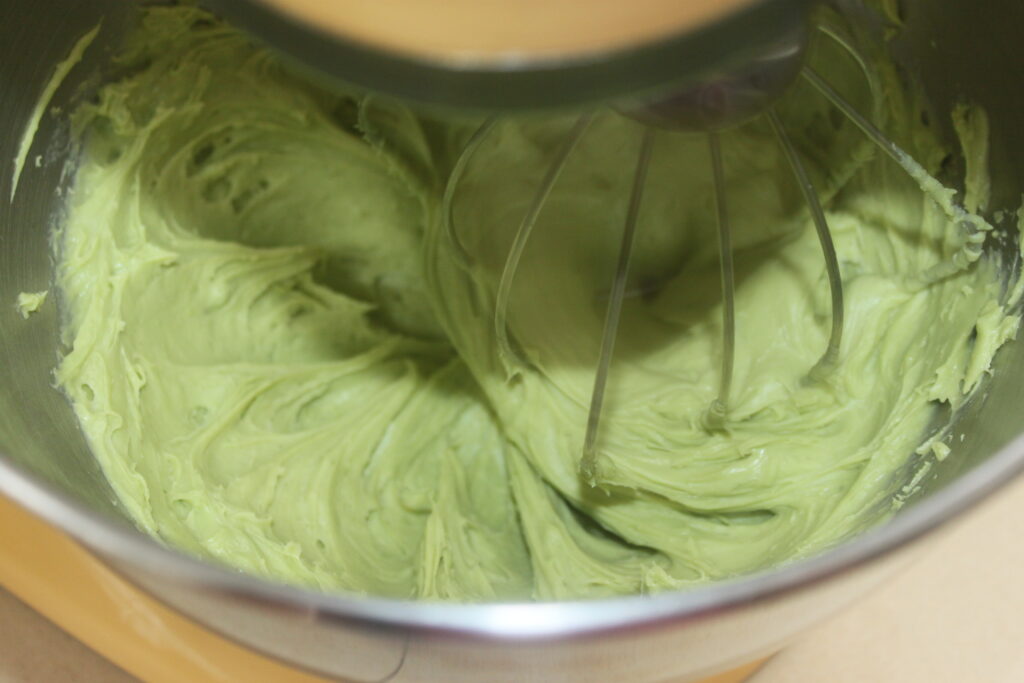 Un bol de batteur sur socle contient de la pâte verte mélangée avec un fouet, parfait pour créer une délicieuse crème basilic chocolat blanc.