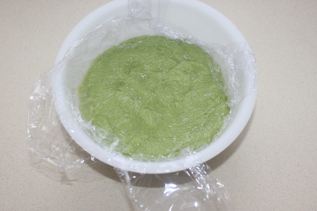 Un bol blanc contenant de la pâte verte recouverte d'une pellicule plastique repose sur un plan de travail de couleur claire, rappelant les ingrédients frais utilisés dans la fabrication des Tartelettes fraise.