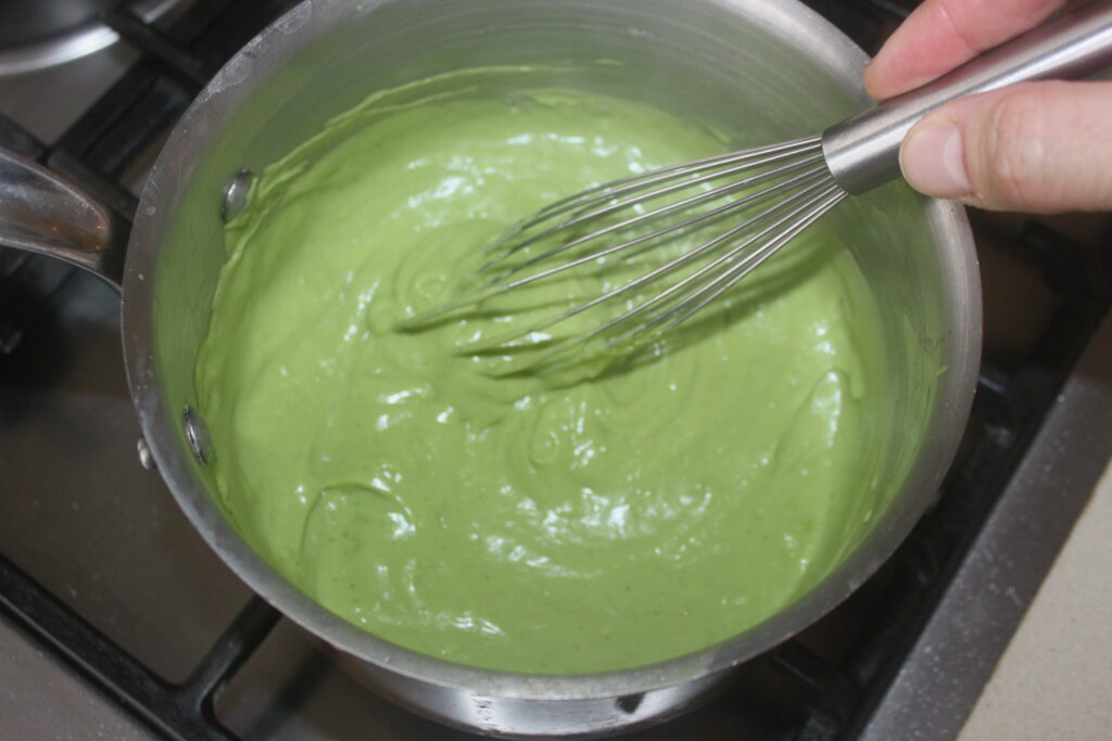 Une personne fouette un mélange vert et épais dans une casserole sur la cuisinière, préparant peut être une délicieuse crème basilic pour accompagner ses Tartelettes fraîches.