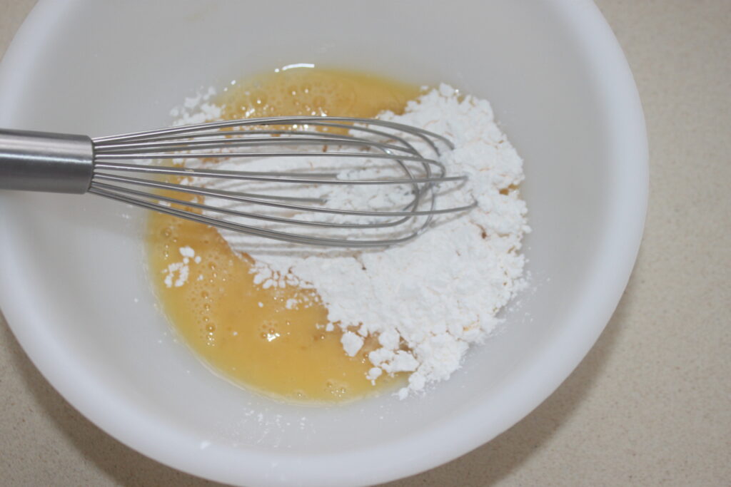 Un bol blanc contient des œufs fêlés et de la farine, avec un fouet en métal placé dessus, prêt à être mélangé : des ingrédients parfaits pour créer de délicieuses Tartelettes fraise.
