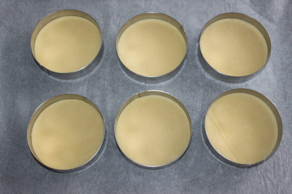 Six pâtons circulaires jaunes nichés dans des emporte pièces en métal disposés sur une plaque à pâtisserie, parfaits pour créer de délicieuses tartelettes fraîches au mascarpone chantilly.