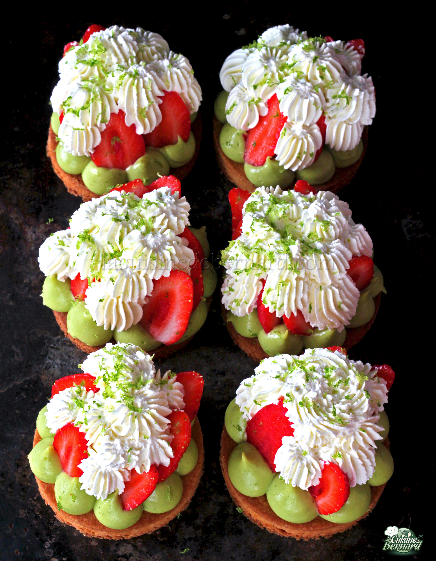 Six petites tartelettes fraise garnies de cuillerées de chantilly mascarpone citron vert, de tranches de fraises et de crème basilic chocolat blanc. Décoré de zestes de citron vert sur fond sombre.