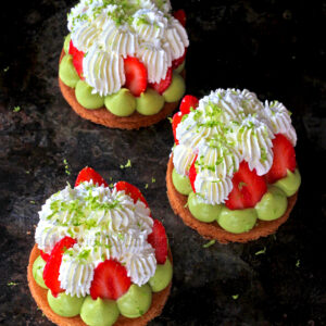Trois Tartelettes fraîches individuelles garnies de mascarpone chantilly, de fraises, de crème verte et de zeste de citron vert sur une surface sombre.