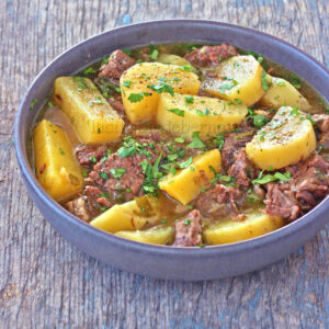 Un bol de ragoût de bœuf avec pommes de terre et garni de persil haché, servi sur une surface en bois.