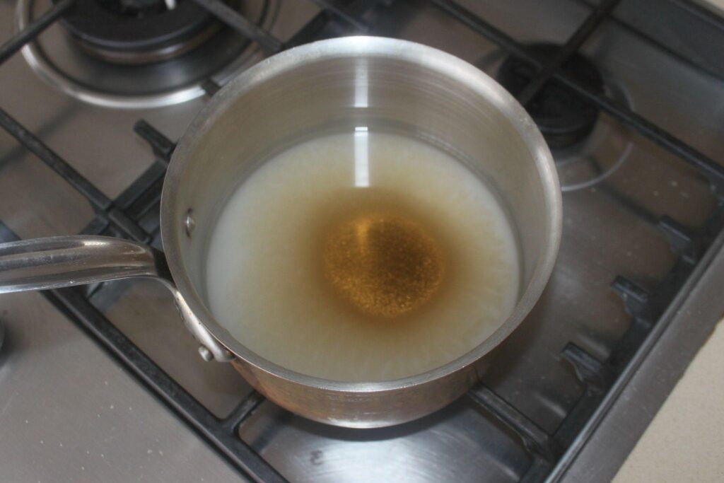 Une casserole avec de l'eau et du sucre cristallisé est posée sur une cuisinière à gaz, le sucre commençant à se dissoudre au centre.