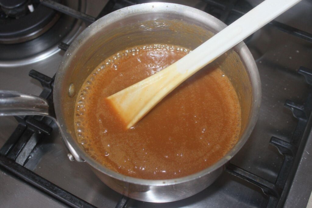 Une casserole avec une sauce au caramel brun remuée par une spatule blanche sur une cuisinière à gaz.