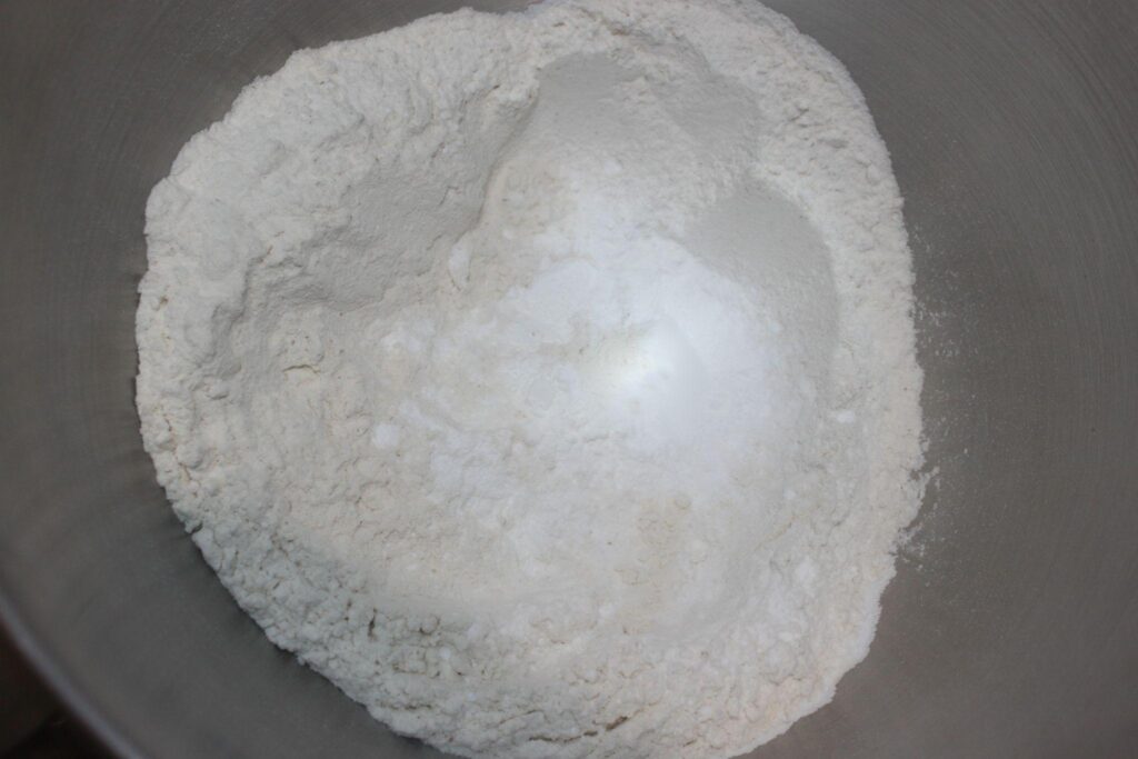 Un bol en métal contenant un monticule d'ingrédients secs blancs et poudreux, probablement de la farine et du sucre, prêts à être mélangés.