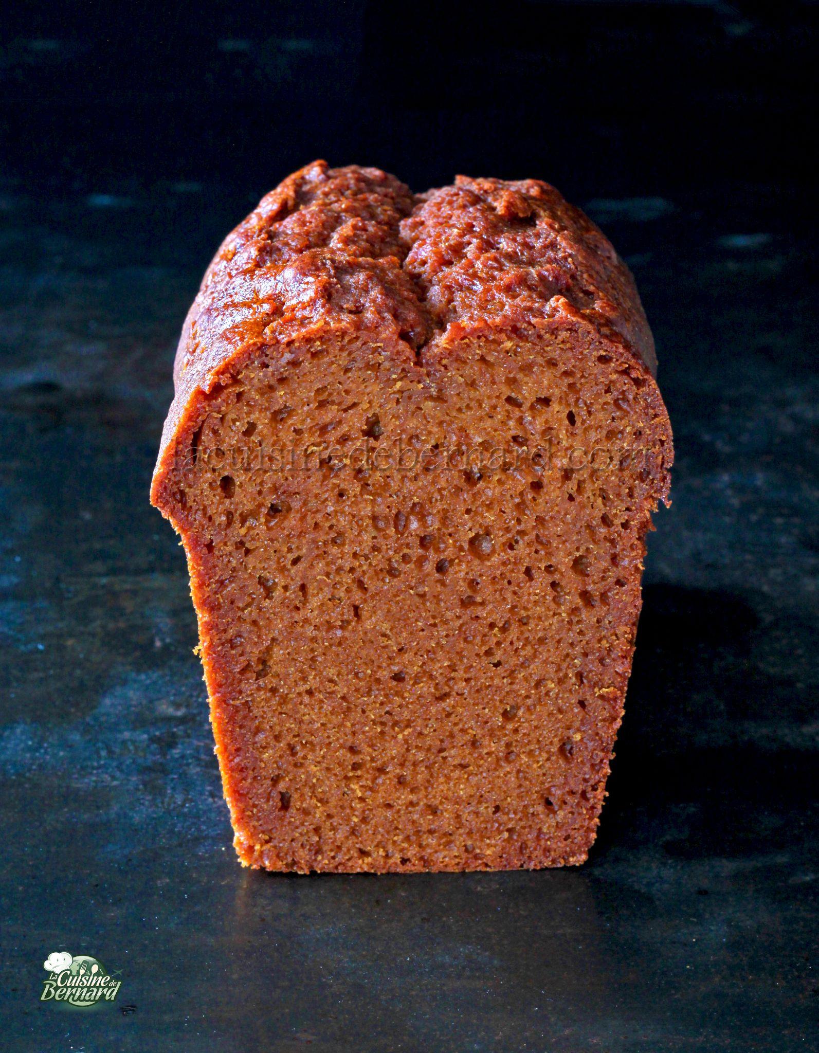 Un pain d'épices se dresse debout sur une surface sombre, montrant sa texture de mie brune, humide et dense.