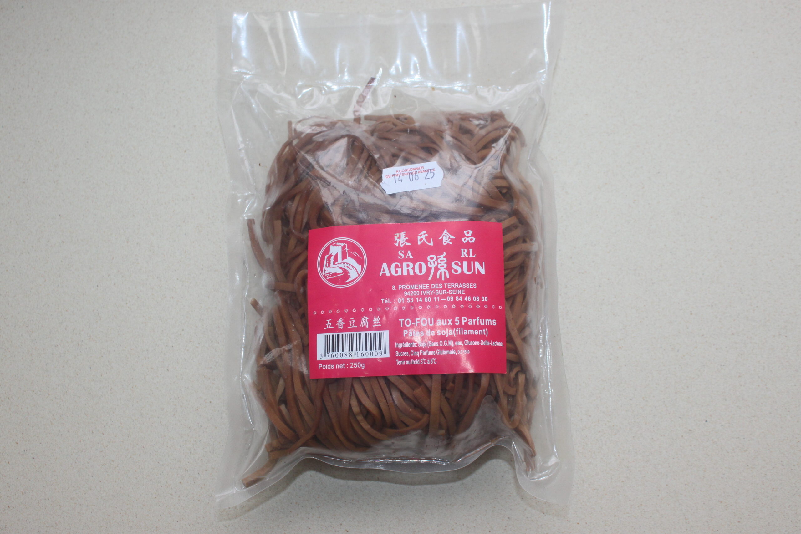 Un emballage en plastique scellé de nouilles de tofu de marque Agro Sun avec une étiquette rouge, marquée Brouillon auto, repose sur une surface de couleur claire.