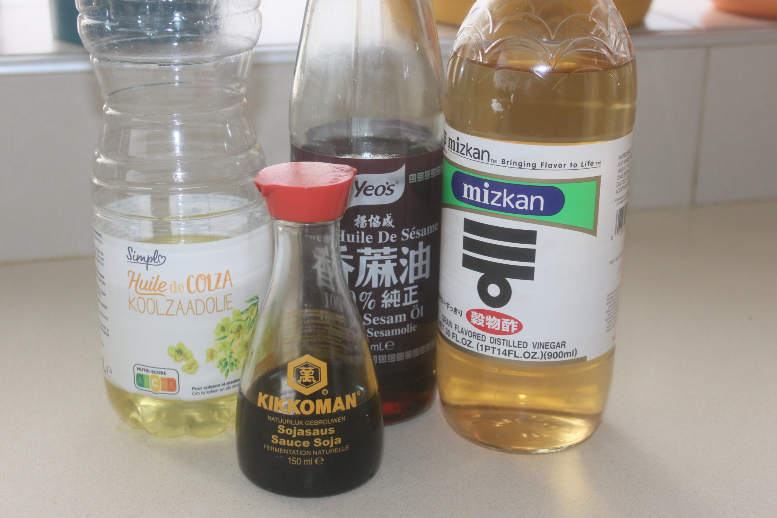 Quatre bouteilles d'ingrédients de cuisine, dont de l'huile de canola, de l'huile de sésame, de la sauce soja et du vinaigre de riz, sont soigneusement disposées sur un comptoir de cuisine de couleur claire à côté d'une machine à Brouillon.